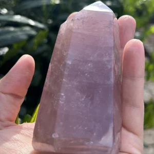 Hand hält Rosenquarz Turm vor grünem Laub und hellem Licht